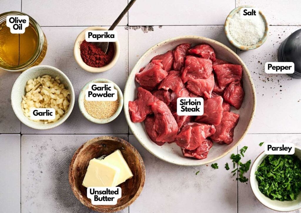Bowls containing ingredients for Air Fryer Garlic Butter Steak Bites—chopped garlic, olive oil, paprika, garlic powder, sirloin steak pieces, unsalted butter, salt, pepper, and chopped parsley—are arranged on a countertop.