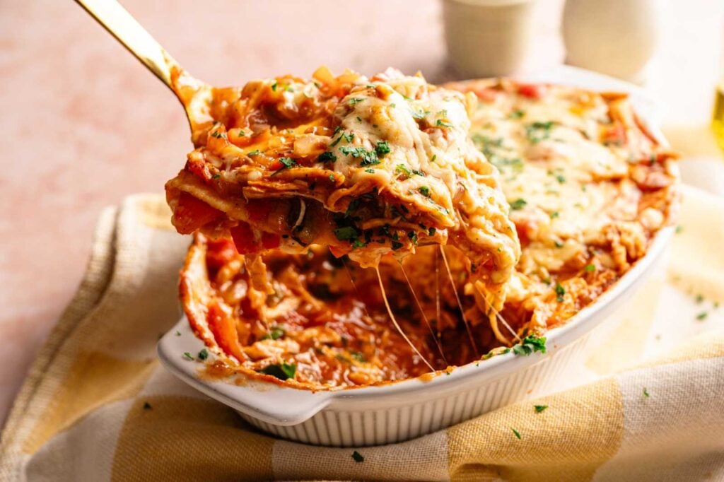 A serving of cheesy King Ranch Casserole is being lifted from a white dish, revealing layers of pasta, savory sauce, and melted cheese.
