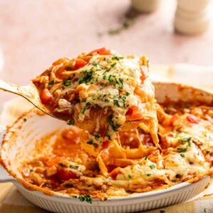 A serving spoon lifts a cheesy King Ranch Casserole from a dish topped with melted cheese and chopped herbs, with a checkered towel underneath.