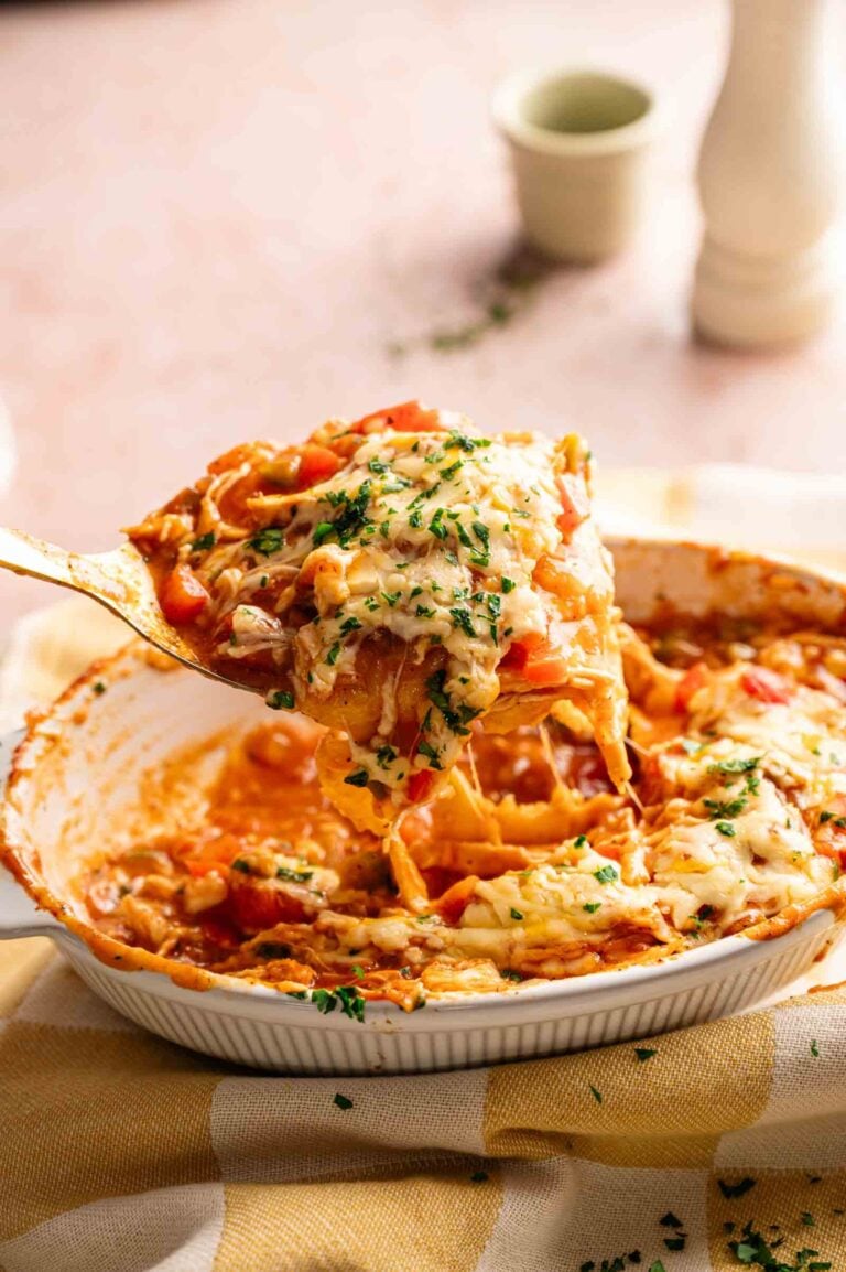 A serving spoon lifts a cheesy King Ranch Casserole from a dish topped with melted cheese and chopped herbs, with a checkered towel underneath.