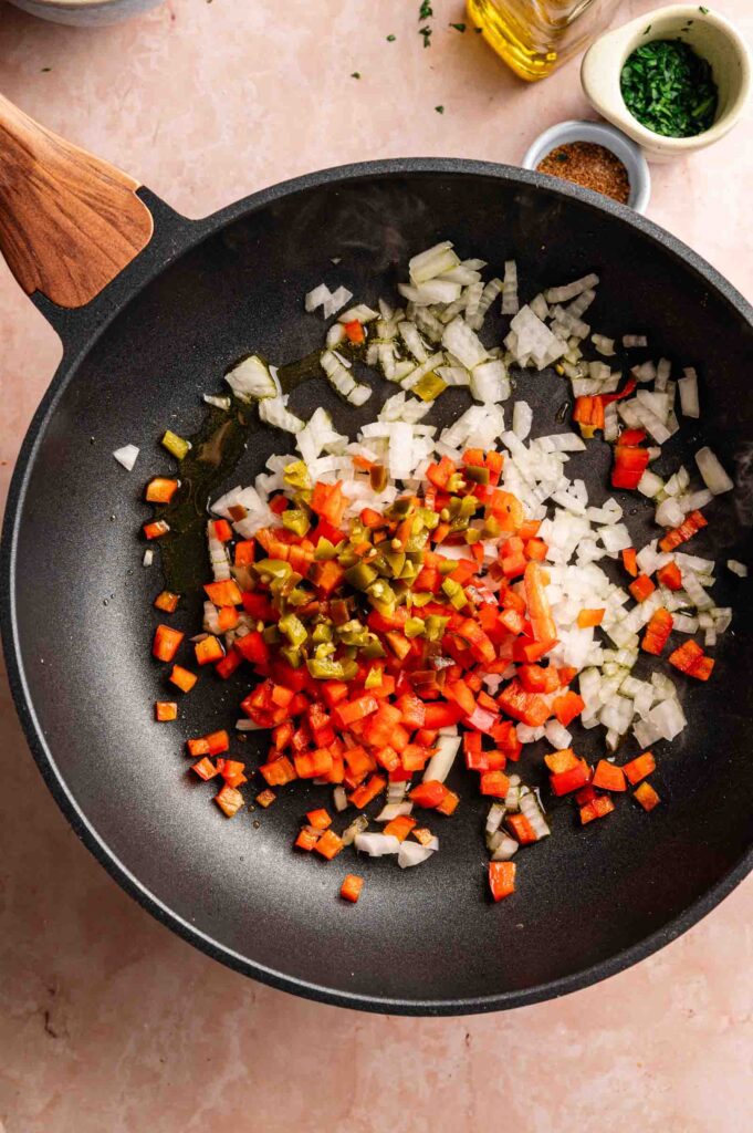Chopped onions, red bell peppers, and green chiles are in a black skillet on a light countertop, ready to be cooked for a classic King Ranch Casserole.