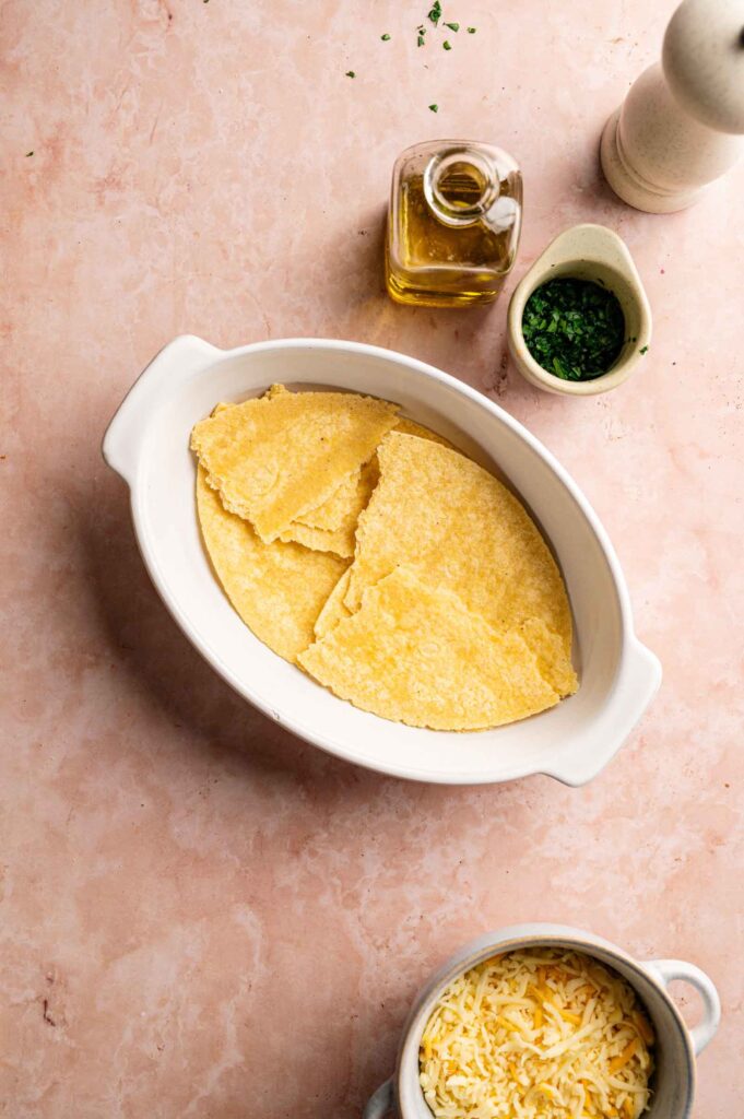 Oval white dish with three corn tortillas, next to a small bowl of shredded cheese, a jug of olive oil, a small bowl of chopped herbs, and a white pepper grinder on a pink surface—perfect fixings for assembling your favorite King Ranch Casserole.