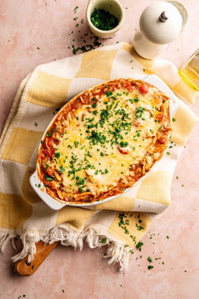 King Ranch Casserole baked to perfection, topped with chopped parsley in a white dish, set on a yellow and white checkered cloth with a pepper grinder and small bowl of herbs nearby.