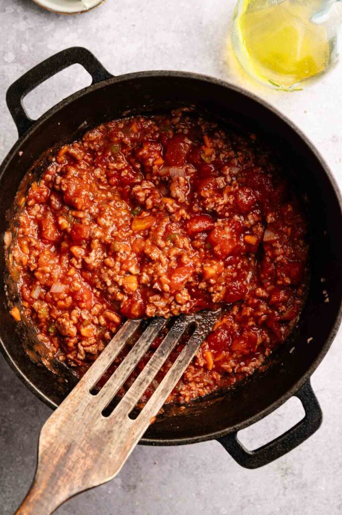 A pot of rich, tomato-based Lamb Bolognese sauce with visible ground meat and a wooden spatula rests on a light countertop next to a partially visible bottle.