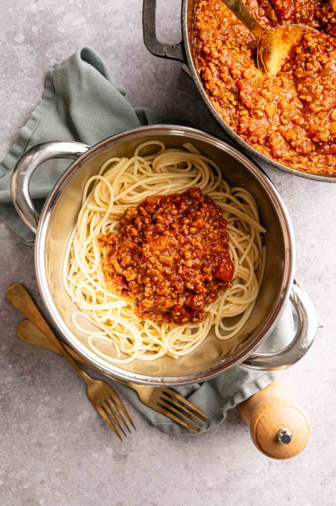 A pot of cooked spaghetti topped with savory Lamb Bolognese sits on a green cloth, with gold utensils and a pepper mill beside it. Another pot containing more rich meat sauce is nearby.