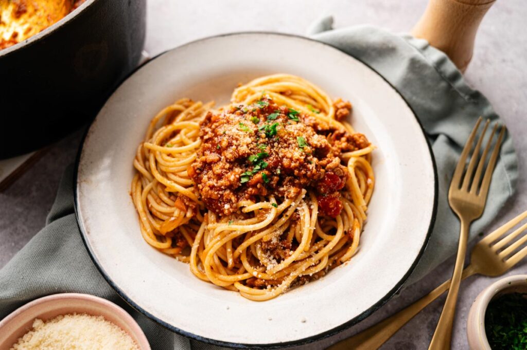 A plate of spaghetti topped with rich Lamb Bolognese and grated cheese, garnished with parsley, sits on a table next to a fork, knife, and a bowl of extra grated cheese.