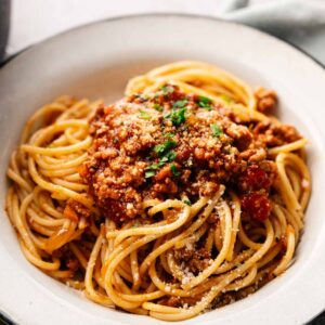 A bowl of spaghetti topped with rich Lamb Bolognese, grated cheese, and chopped herbs, served on a white plate.