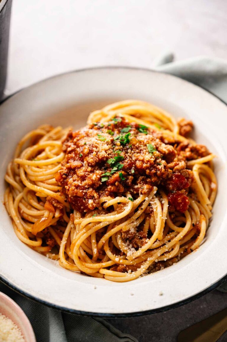 A bowl of spaghetti topped with rich Lamb Bolognese, grated cheese, and chopped herbs, served on a white plate.