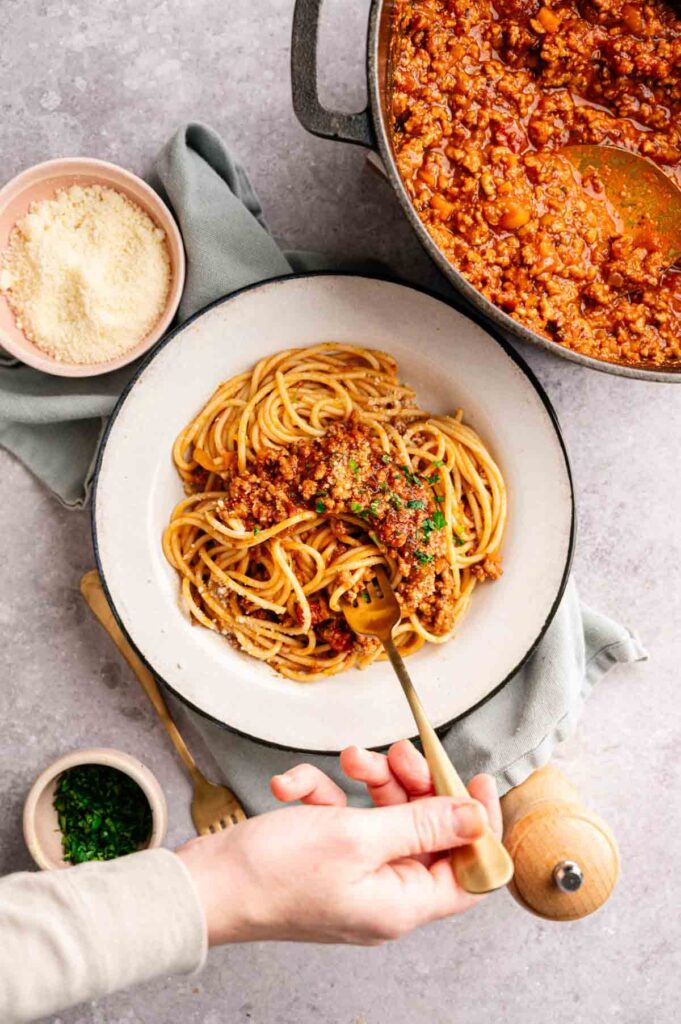 A bowl of spaghetti topped with rich Lamb Bolognese sits on a table next to a pot of sauce, grated cheese, chopped herbs, and a hand holding a fork.