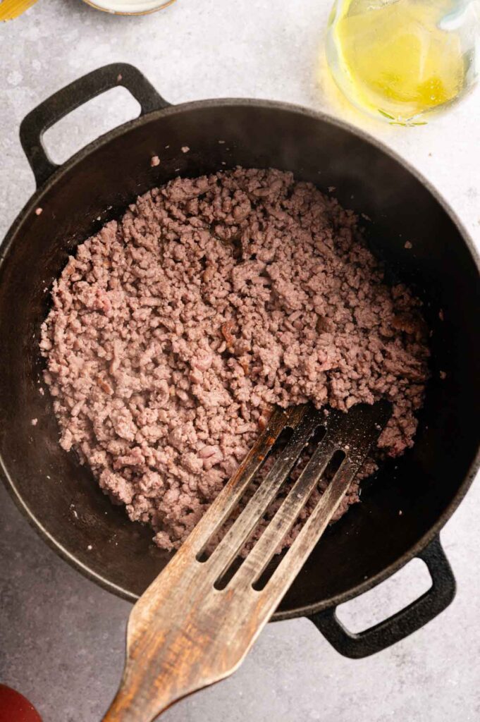 Ground beef is being cooked in a black cast iron pot with a slotted spatula resting inside, creating the perfect base for a hearty Lamb Bolognese.