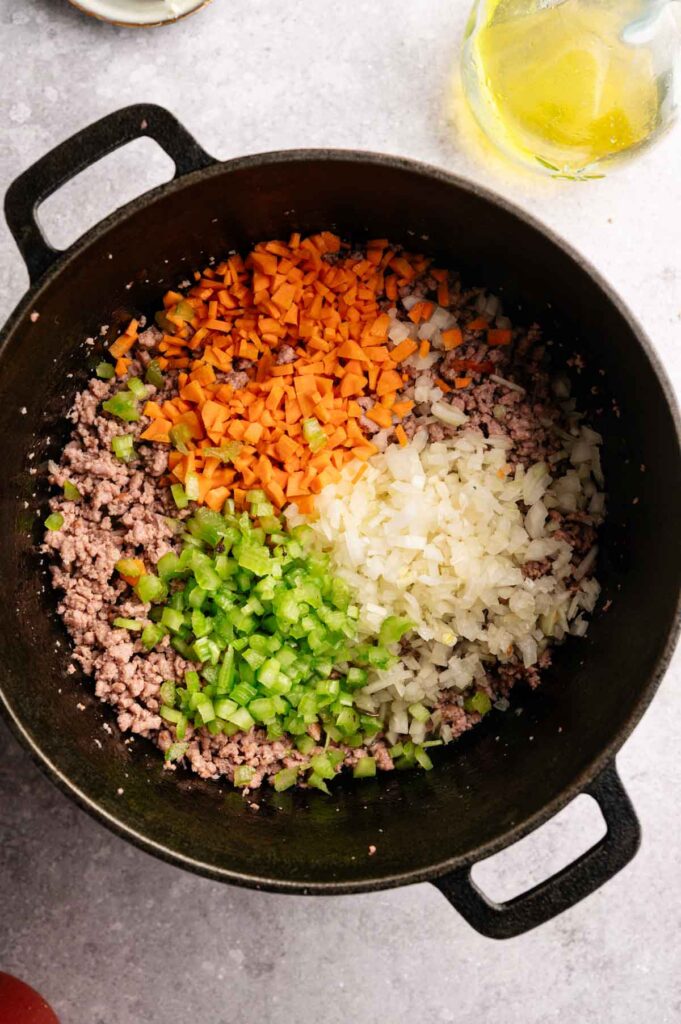 A black pot with ground meat, chopped carrots, onions, and green bell pepper arranged in sections—perfect for starting a hearty Lamb Bolognese, ready to be cooked.