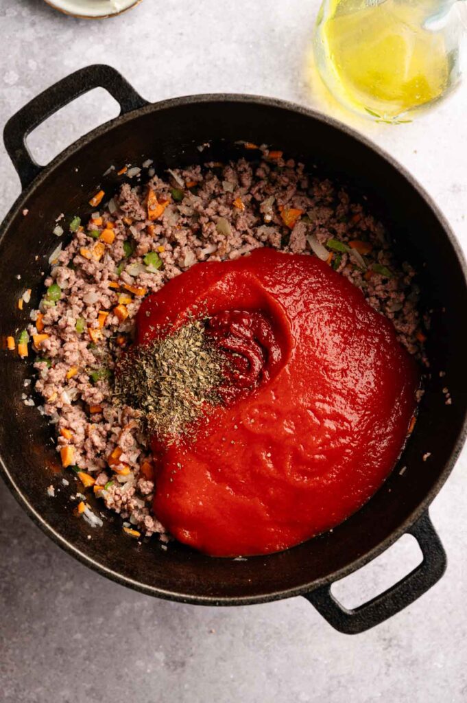A black pot contains cooked ground meat, diced vegetables, and a large portion of tomato sauce with seasonings on top, ready to be mixed into a rich Lamb Bolognese.