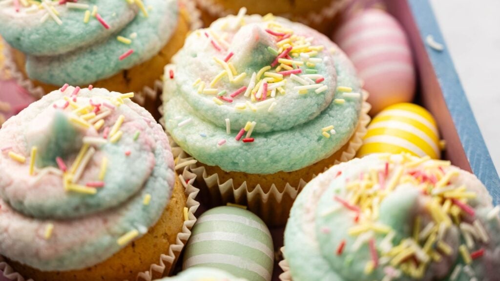 Cupcakes with pastel blue and pink frosting topped with sprinkles, surrounded by pastel-colored striped and solid eggs in a box.