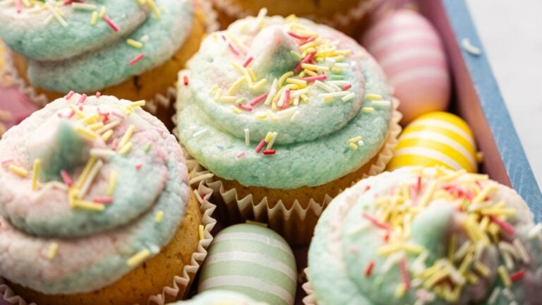 Cupcakes with pastel blue and pink frosting topped with sprinkles, surrounded by pastel-colored striped and solid eggs in a box.