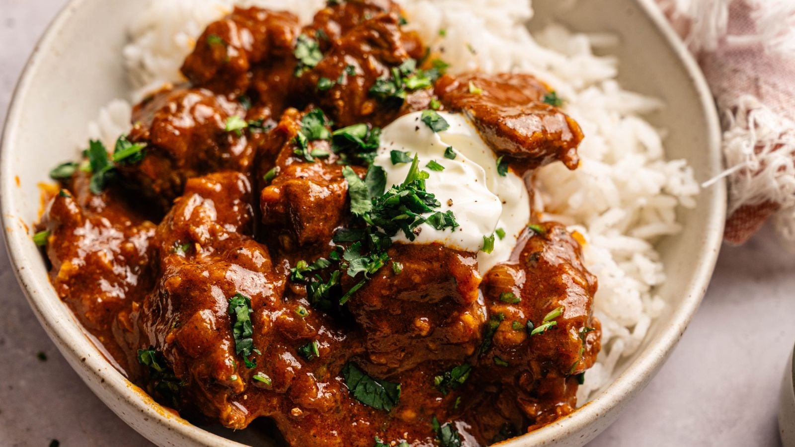A bowl of white rice topped with chunks of beef in a reddish-brown sauce, garnished with fresh herbs and a dollop of cream.