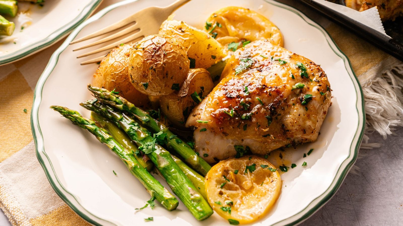 A plate with roasted chicken breast, golden potatoes, asparagus spears, and lemon slices, garnished with chopped herbs.