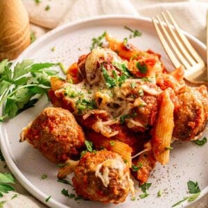 A plate of Meatball Casserole featuring baked pasta with marinara sauce, melted cheese, and hearty meatballs, garnished with chopped parsley, sits next to a fork and fresh parsley sprigs.