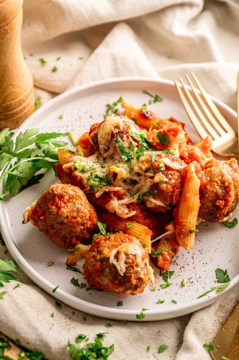 A plate of Meatball Casserole featuring baked pasta with marinara sauce, melted cheese, and hearty meatballs, garnished with chopped parsley, sits next to a fork and fresh parsley sprigs.