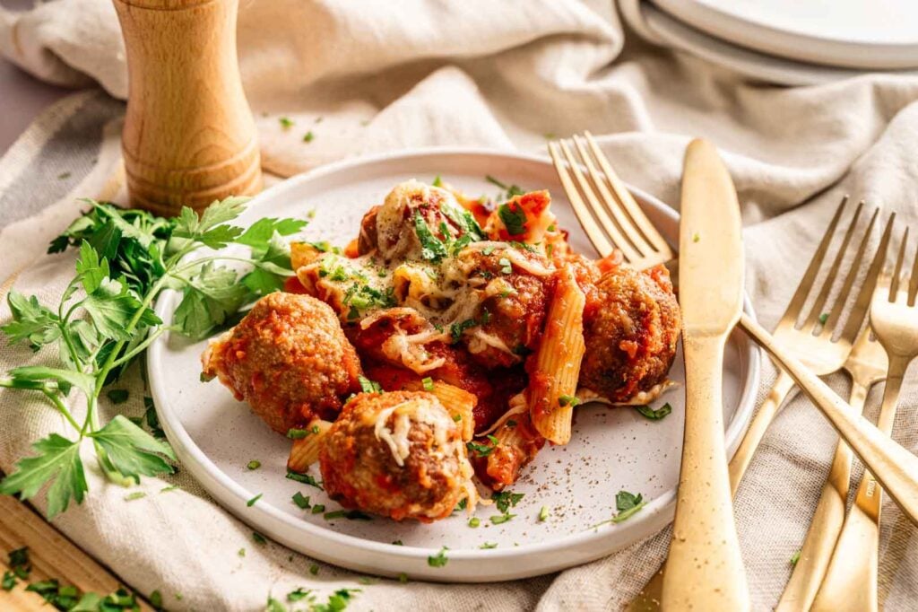 A plate of Meatball Casserole featuring baked penne pasta with tomato sauce, melted cheese, and juicy meatballs, garnished with chopped parsley and served with gold-colored cutlery.
