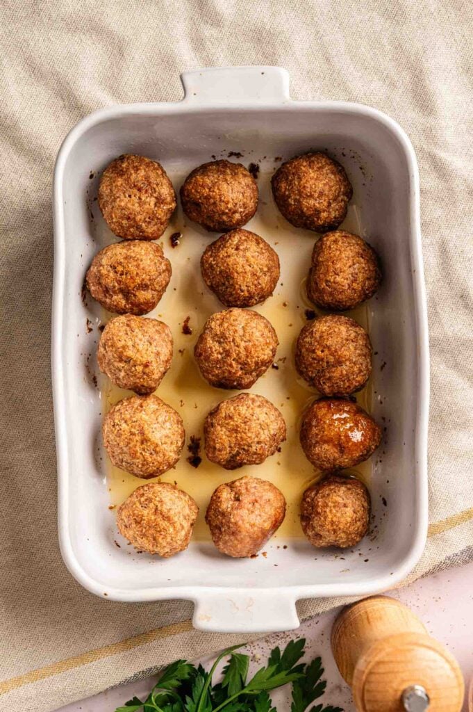 A white baking dish containing a savory Meatball Casserole with twelve cooked meatballs and flavorful juices pooled at the bottom, placed on a light cloth surface.