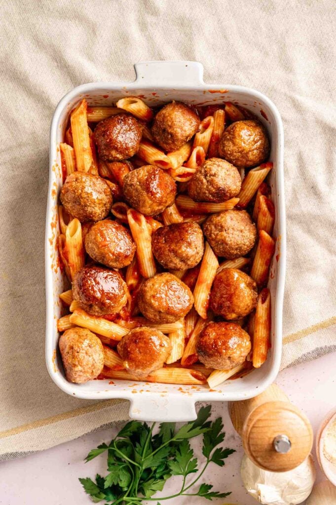 Meatball Casserole featuring baked meatballs and penne pasta in tomato sauce is arranged in a white rectangular baking dish on a neutral cloth.