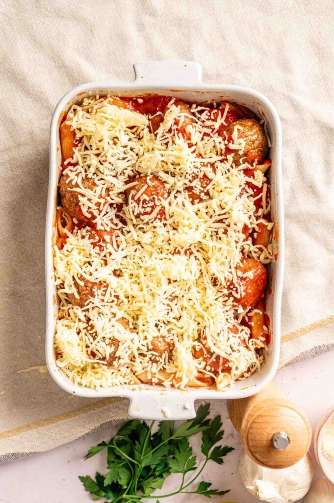 A rectangular white baking dish filled with Meatball Casserole&mdash;pasta, meatballs, tomato sauce, and shredded cheese&mdash;ready to be baked. A pepper grinder and parsley are nearby.