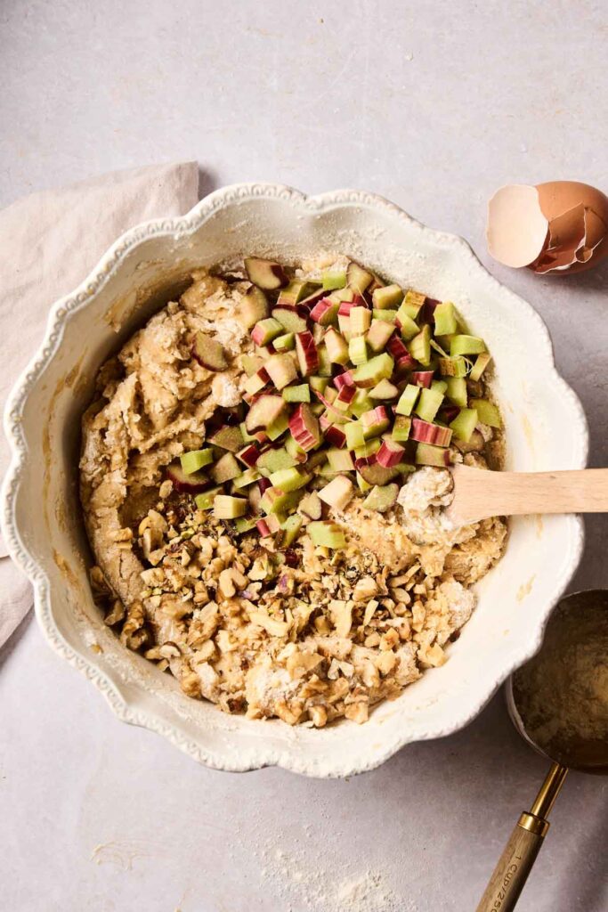 A mixing bowl with batter for Rhubarb Muffins, chopped rhubarb, and walnuts, a wooden spoon inside, a cracked eggshell, and measuring cup nearby on a light surface.