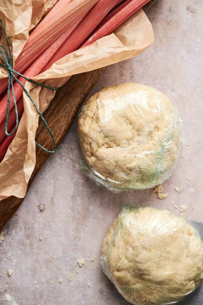 Two balls of dough wrapped in plastic wrap sit on a countertop beside a bundle of rhubarb stalks tied with twine and wrapped in parchment paper—perfectly prepared for making a homemade Air Fryer Rhubarb Pie.