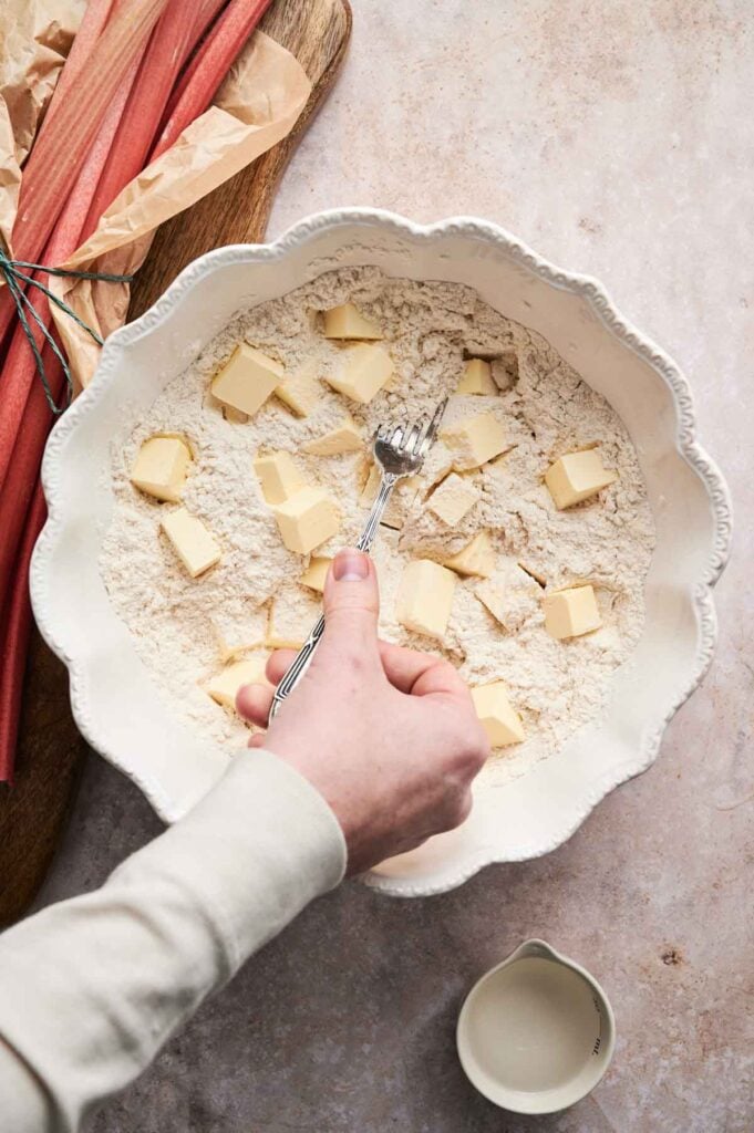 A hand uses a pastry blender to cut butter into flour in a white mixing bowl, preparing the base for an Air Fryer Rhubarb Pie, with fresh rhubarb stalks and a small jug nearby on a light surface.