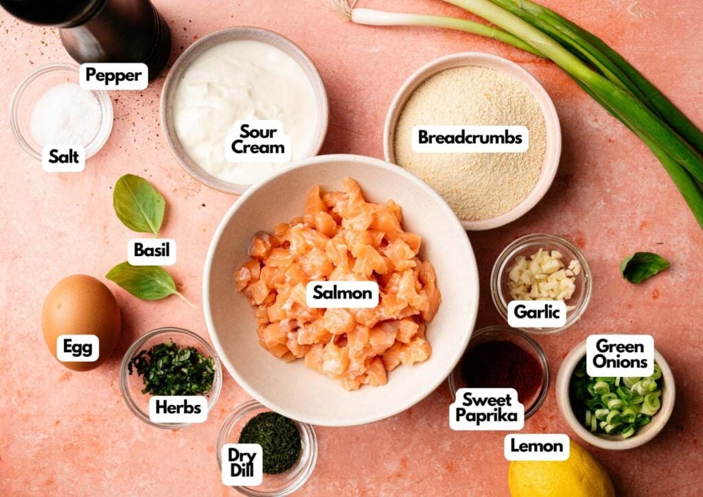 Ingredients for Air Fryer Salmon Patties arranged on a pink surface, including diced salmon, sour cream, breadcrumbs, garlic, green onions, lemon, egg, salt, pepper, basil, herbs, sweet paprika, and dry dill.