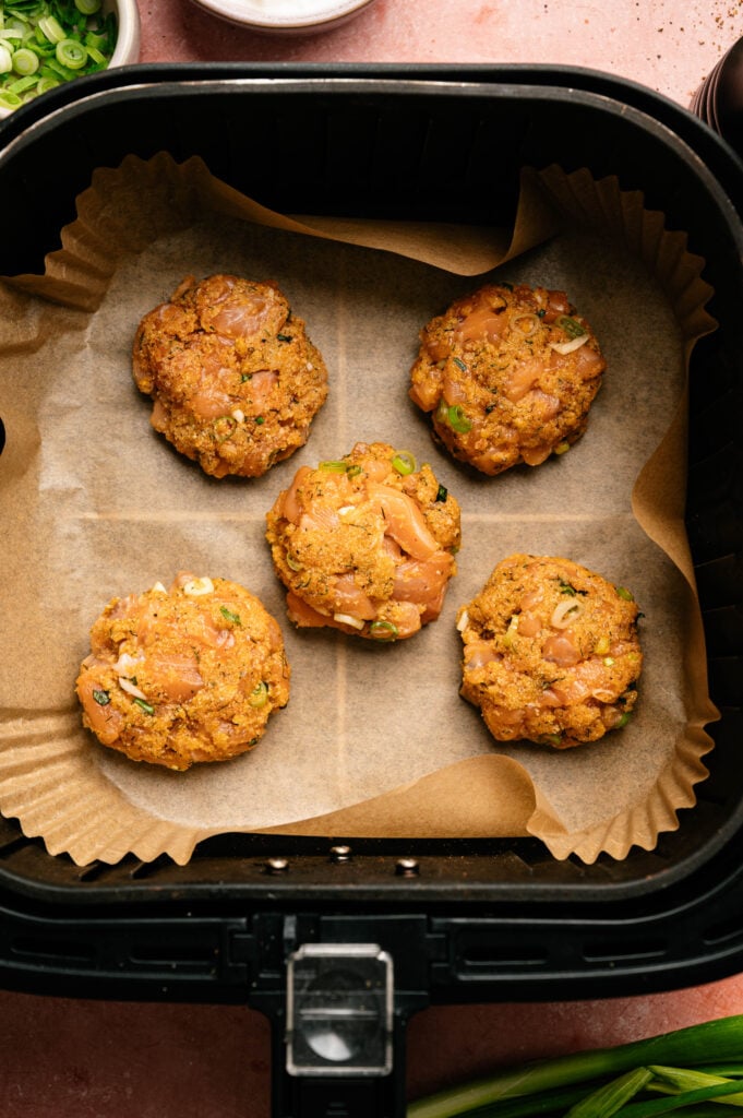 Five raw chicken patties with herbs are arranged on parchment paper inside an air fryer basket, ready to be cooked&mdash;perfect for those who also enjoy making Air Fryer Salmon Patties at home.