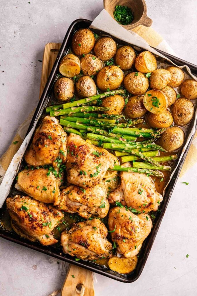 A sheet pan of chicken thighs with roasted asparagus and halved baby potatoes, all garnished with chopped herbs.