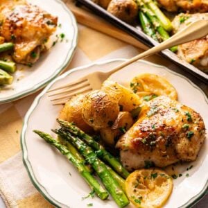 A plate with Sheet Pan Chicken Thighs, asparagus, lemon slices, and roasted potatoes sits beside a fork. In the background, a tray holds more of the flavorful meal.