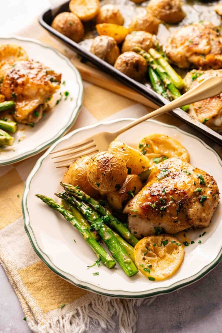 A plate with Sheet Pan Chicken Thighs, asparagus, lemon slices, and roasted potatoes sits beside a fork. In the background, a tray holds more of the flavorful meal.