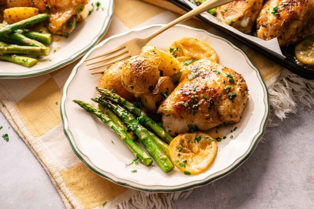 A plate of Sheet Pan Chicken Thighs with roasted baby potatoes, asparagus, and lemon slices, garnished with chopped herbs, served on a white plate with a gold fork.