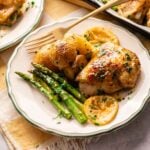 A plate of Sheet Pan Chicken Thighs with roasted baby potatoes, asparagus, and lemon slices, garnished with chopped herbs, served on a white plate with a gold fork.