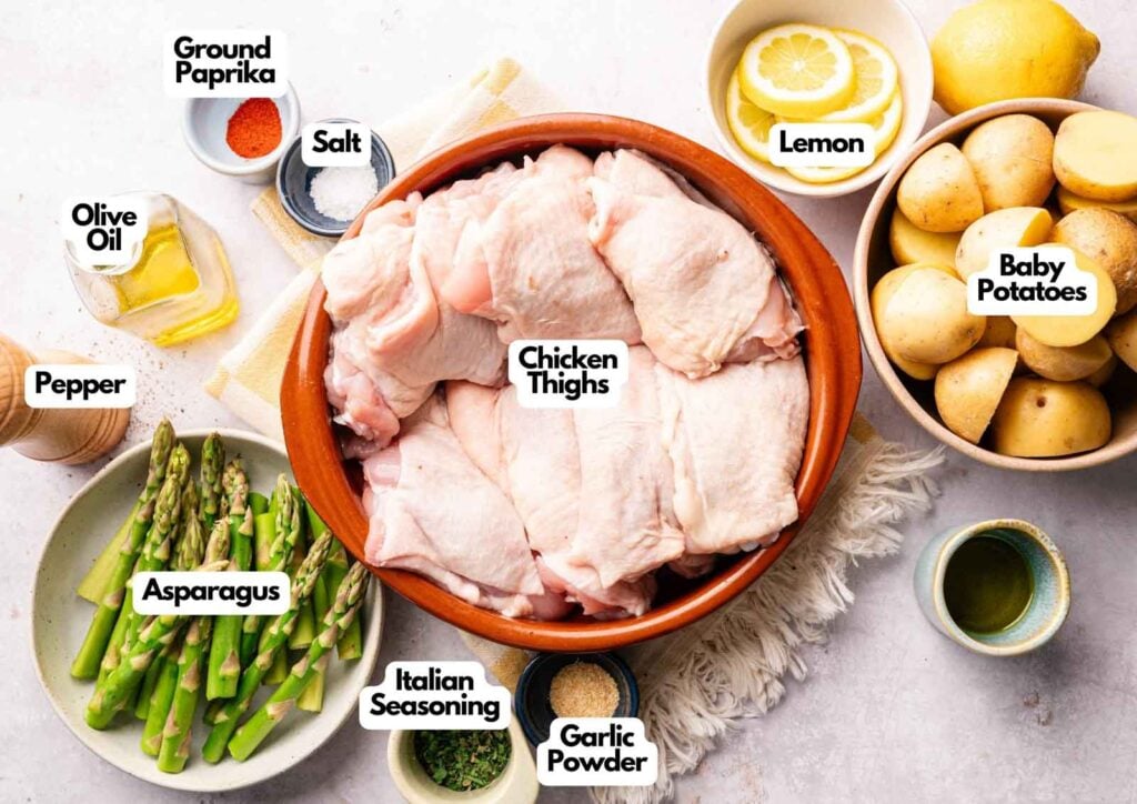 A bowl of raw chicken thighs, perfect for making Sheet Pan Chicken Thighs, is surrounded by labeled ingredients: ground paprika, salt, lemon, baby potatoes, olive oil, pepper, asparagus, Italian seasoning, and garlic powder.