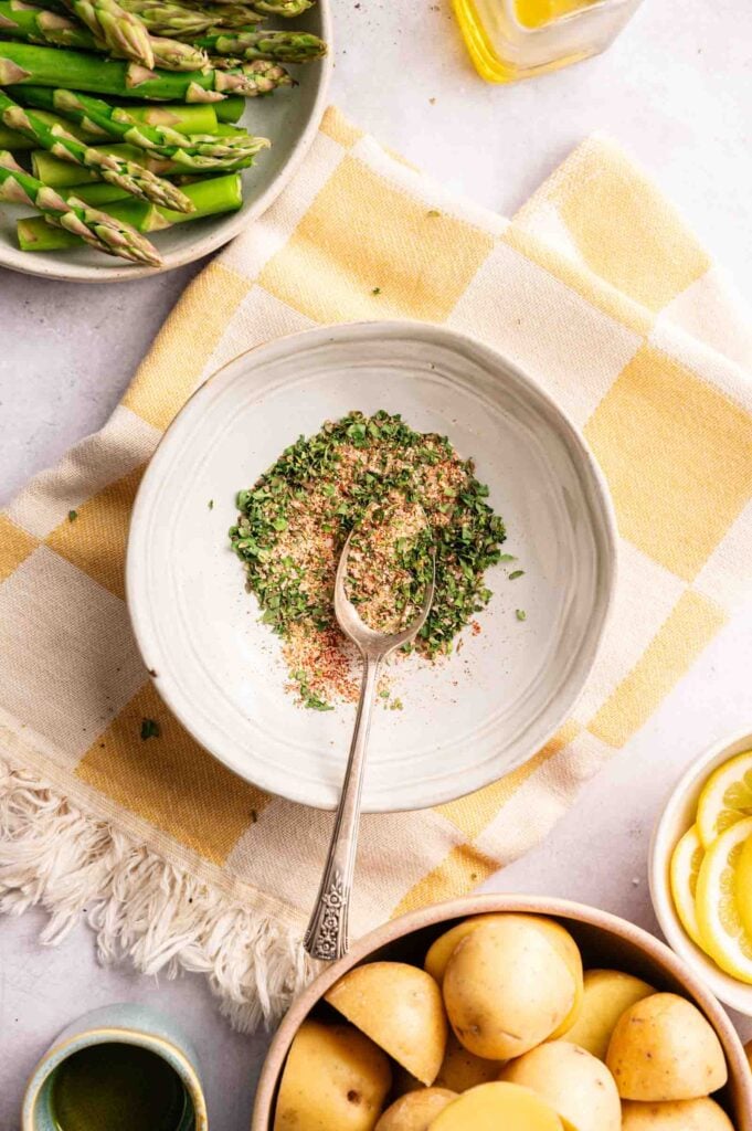 A bowl with a spoon rests on a yellow checkered cloth, holding a blend of dried herbs and seasonings—perfect for preparing delicious Sheet Pan Chicken Thighs. Surrounding items include asparagus, potatoes, lemon slices, and a bottle of olive oil.