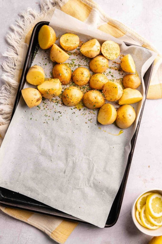 Halved baby potatoes sprinkled with herbs and seasoning are arranged on a parchment-lined baking sheet, perfect for roasting alongside juicy Sheet Pan Chicken Thighs. A bowl with lemon slices is nearby on a yellow and white striped cloth.