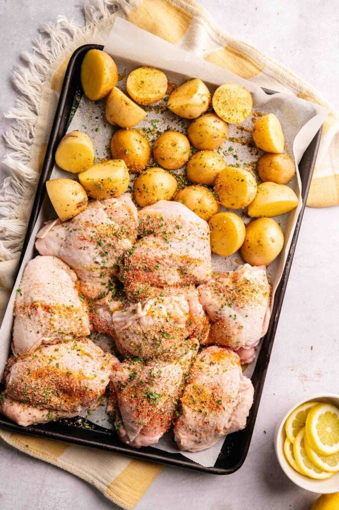 Sheet Pan Chicken Thighs and halved potatoes, seasoned with herbs and spices, are arranged on a parchment-lined baking sheet, ready to be cooked.