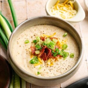 A bowl of creamy Slow Cooker Potato Soup topped with shredded cheese, bacon, and chopped green onions sits on a tiled surface next to fresh green onions and a small bowl of cheese.