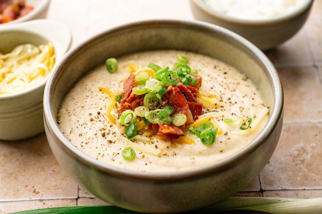 A bowl of creamy Slow Cooker Potato Soup topped with shredded cheese, bacon pieces, sliced green onions, and cracked black pepper.