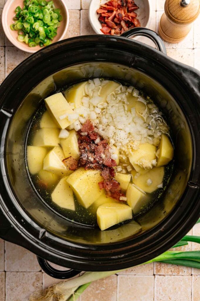 Ingredients for Slow Cooker Potato Soup—cubed potatoes, chopped onions, minced garlic, bacon, and broth—are ready in a slow cooker; sliced green onions and crumbled bacon sit in small bowls nearby.