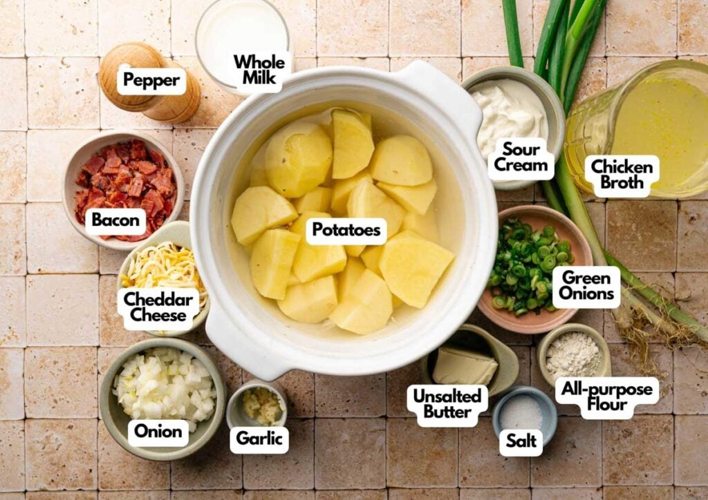 Ingredients for Slow Cooker Potato Soup, including potatoes, bacon, cheddar cheese, onion, garlic, pepper, milk, sour cream, chicken broth, green onions, butter, flour, and salt are laid out on a tiled surface.