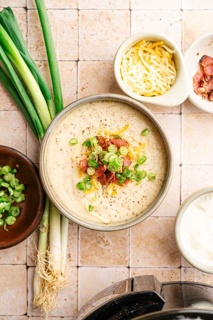 A bowl of creamy Slow Cooker Potato Soup topped with shredded cheese, bacon, and chopped green onions, surrounded by fresh green onions and bowls of toppings on a tiled surface.