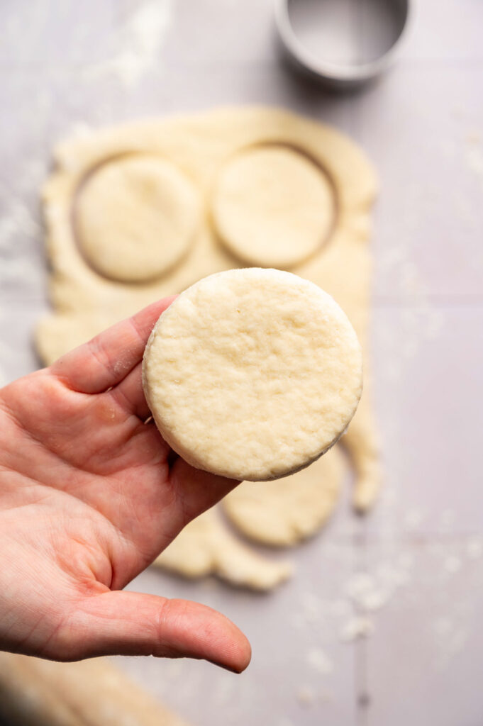 A hand holds a round, uncooked Air Fryer Sourdough Biscuits dough cutout; more cutouts and flour are visible on a flat surface in the background.