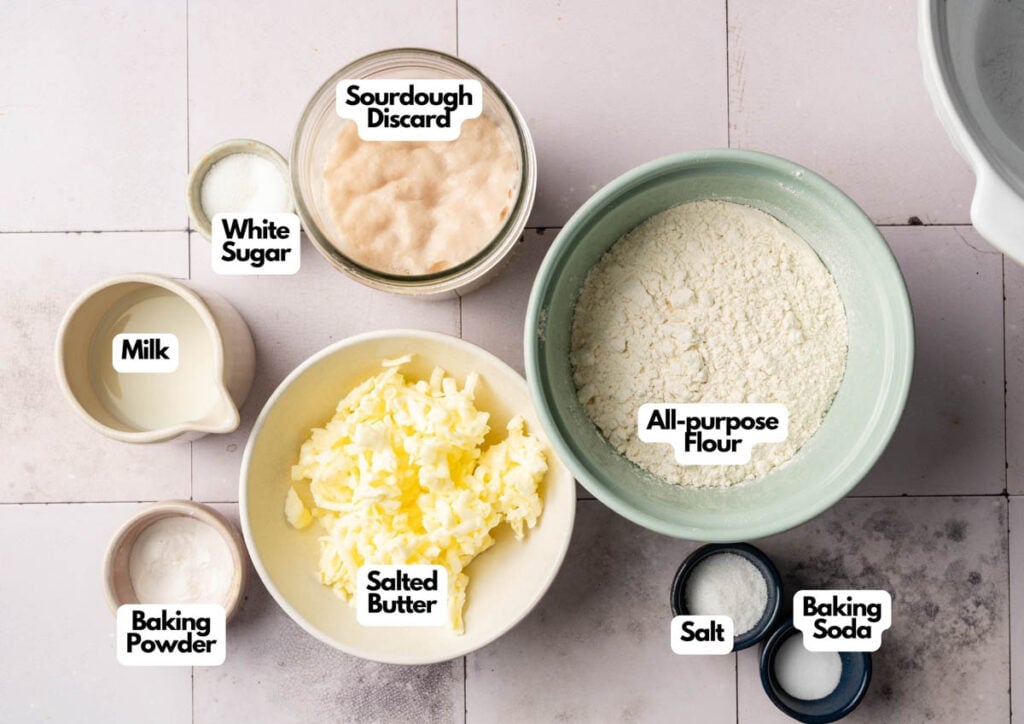Top-down view of labeled baking ingredients for Air Fryer Sourdough Biscuits, including sourdough discard, white sugar, milk, baking powder, salted butter, all-purpose flour, salt, and baking soda on a light surface.