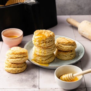 Three stacks of Air Fryer Sourdough Biscuits on a plate and table, drizzled with honey, with a bowl of honey, a honey dipper, a brush, and an air fryer in the background.