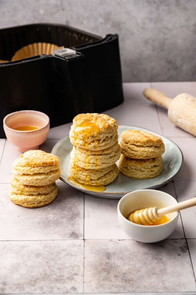 Three stacks of Air Fryer Sourdough Biscuits on a plate and table, drizzled with honey, with a bowl of honey, a honey dipper, a brush, and an air fryer in the background.