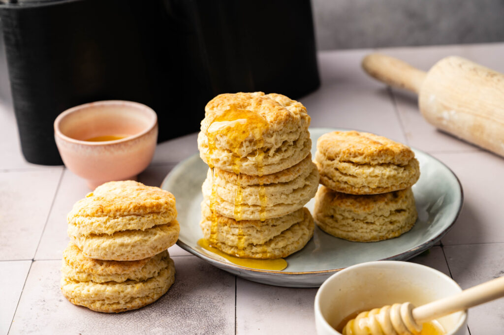 A plate of stacked Air Fryer Sourdough Biscuits drizzled with honey sits on a tiled surface, surrounded by a honey dipper, bowl, rolling pin, and a small cup of honey.
